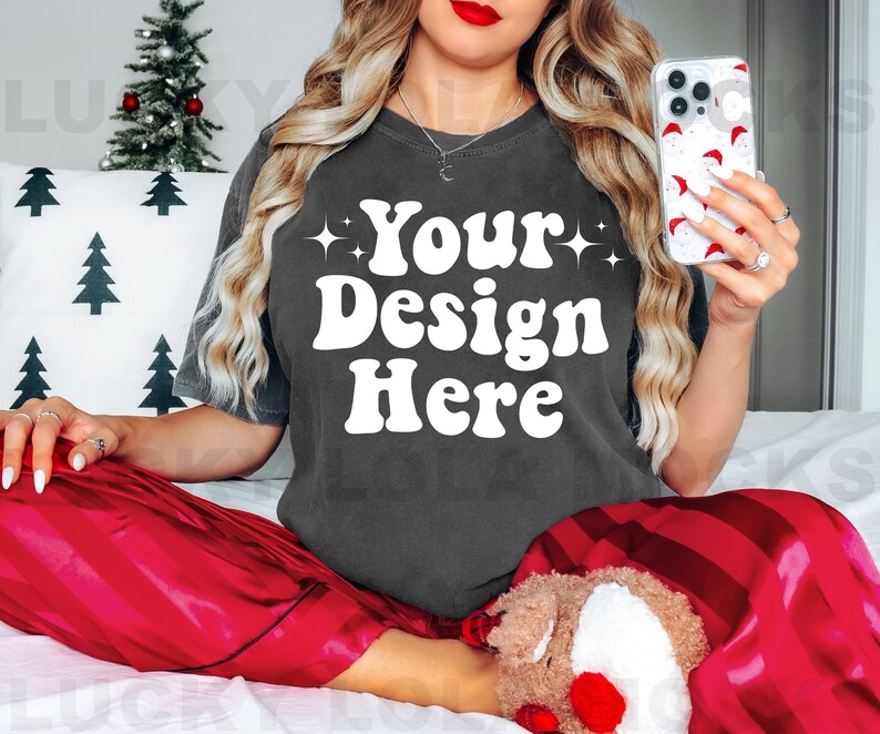 May include: A woman wearing a dark gray t-shirt with the text "Your Design Here" in white. She is sitting on a white bed with red and white striped pajamas. She is holding a clear phone case with a red and white pattern.