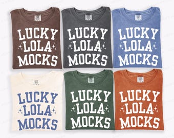 May include: Six t-shirts in various colors, including brown, gray, blue, cream, green, and orange. Each shirt displays the text "LUCKY LOLA MOCKS" in a bold, white font. The shirts are neatly folded, showcasing the design.
