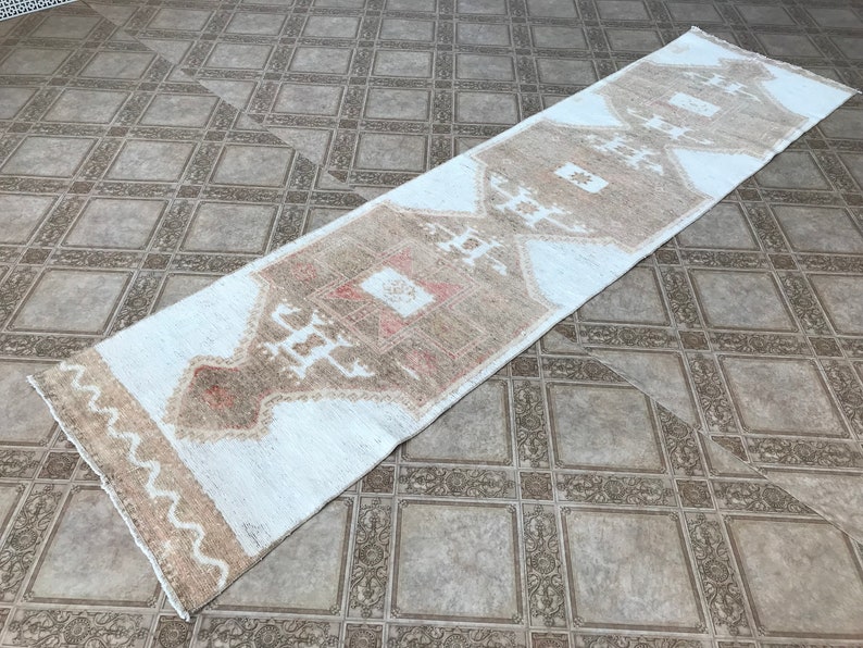 May include: A white and beige runner rug with a geometric pattern. The rug has a distressed look and is made of a soft, woven material.