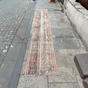 May include: A long, narrow rug with a cream background and a repeating pattern of red, blue, and black geometric shapes. The rug is lying on a gray cobblestone street.