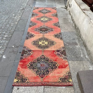 May include: A long, narrow rug with a traditional Persian design. The rug is a mix of red, pink, cream, and black colours. The rug has a fringe on both ends.