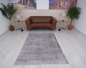 Puede incluir: Una alfombra gris y blanca desgastada con un patrón sutil. La alfombra está colocada sobre un suelo de madera clara en un salón con un sofá de cuero marrón y dos mesas auxiliares.