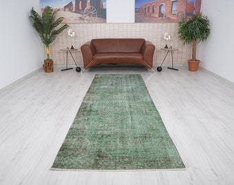 May include: A green vintage rug with a distressed look, laid out on a light wood floor in a room with a brown leather sofa and two potted plants.