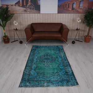 May include: A teal blue vintage rug with a faded floral pattern in the center. The rug is laid out on a light wood floor in a room with a brown leather sofa and two side tables.