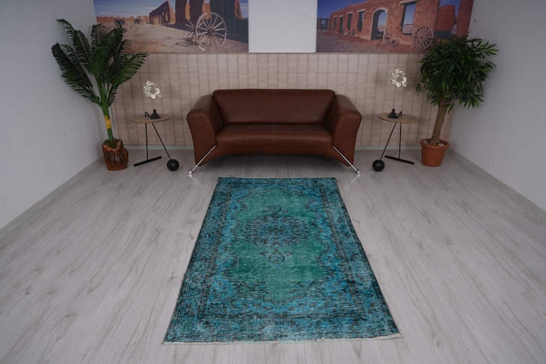 May include: A teal blue vintage rug with a faded floral pattern in the center. The rug is laid out on a light wood floor in a room with a brown leather sofa and two side tables.