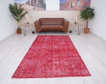 Könnte beinhalten: Ein roter Vintage-Teppich mit einem weißen und grauen Muster. Der Teppich liegt auf einem hellen Holzboden in einem Raum mit einem braunen Ledersofa und zwei Topfpflanzen.