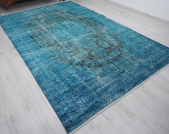 May include: A rectangular, distressed-look rug in shades of blue and teal. The rug features a central, darker pattern with a faded appearance. The rug is placed on a light-colored wooden floor, creating a contrast.