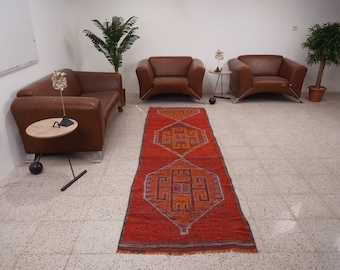 May include: A red, orange, and blue patterned rug with geometric designs. The rug is laid out on a white tiled floor in a room with two brown leather armchairs and a small table.