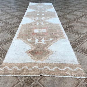 May include: A white runner rug with a beige and pink geometric pattern. The rug has a fringed edge and is made of a soft, woven material.
