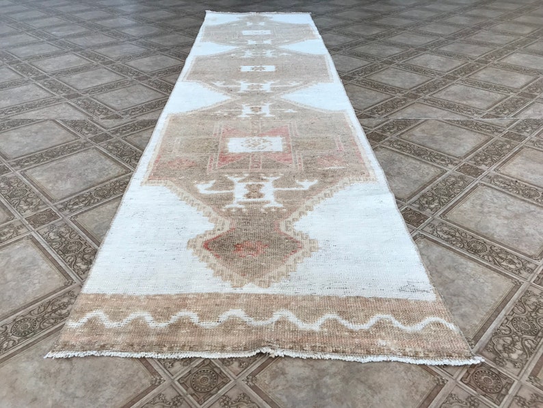 May include: A white runner rug with a beige and pink geometric pattern. The rug has a fringed edge and is made of a soft, woven material.