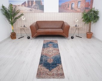 May include: A rectangular, vintage-style rug with a central medallion design. The rug features a dark blue field with intricate patterns in shades of red, beige, and cream. The rug is placed on a light-colored wooden floor, with a brown leather sofa in the background.