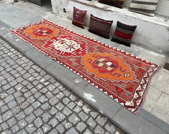 Könnte beinhalten: Ein langer, rechteckiger Vintage-Teppich mit einem geometrischen Muster in Rot-, Orange- und Cremetönen. Der Teppich liegt auf einer Steinoberfläche, mit dekorativen Kissen im Hintergrund.
