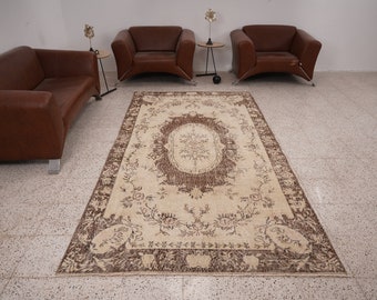 Op de afbeelding: Een vintage crème en bruin kleed met een bloemenpatroon en een centraal medaillon. Het kleed ligt op een betegelde vloer met twee bruine leren stoelen aan weerszijden.