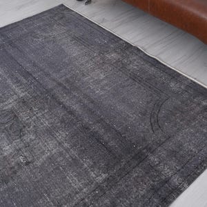 May include: A rectangular, distressed-look rug in shades of gray and white. The rug features a faded, vintage appearance with a subtle border design. The rug is placed on a light-colored wooden floor, with a brown leather sofa visible in the background.