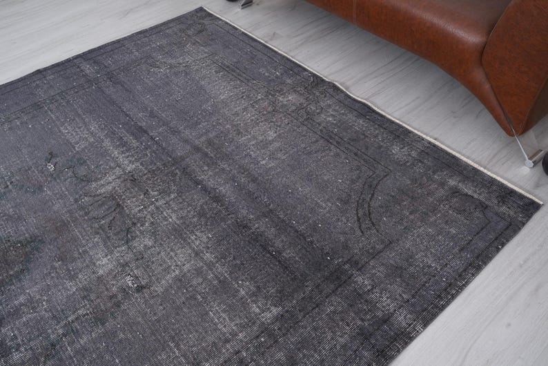 May include: A rectangular, distressed-look rug in shades of gray and white. The rug features a faded, vintage appearance with a subtle border design. The rug is placed on a light-colored wooden floor, with a brown leather sofa visible in the background.