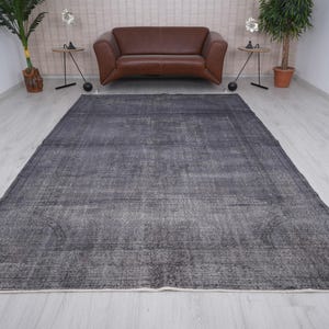 May include: A large, rectangular, dark gray rug with a distressed, vintage appearance. The rug has a faded, mottled pattern and a cream-colored border. The rug is placed on a light-colored wooden floor, with a brown leather sofa and plants in the background.