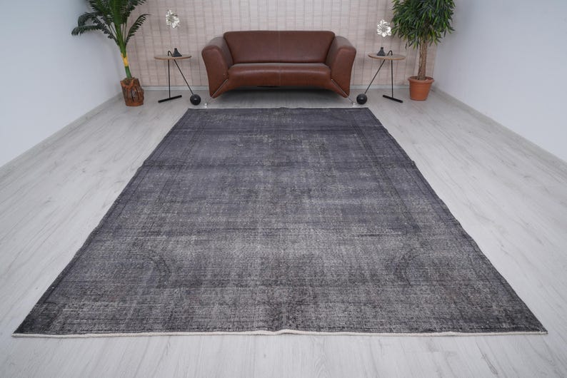 May include: A large, rectangular, dark gray rug with a distressed, vintage appearance. The rug has a faded, mottled pattern and a cream-colored border. The rug is placed on a light-colored wooden floor, with a brown leather sofa and plants in the background.