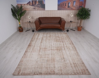 Op de afbeelding: Een lichtbruin vintage tapijt met een verweerde look, met een vervaagd patroon en een subtiele rand. Het tapijt ligt op een lichte houten vloer in een kamer met een bruine leren bank en twee planten in potten.