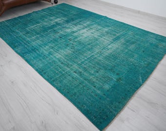 May include: A rectangular, distressed turquoise rug with a faded appearance. The rug has a textured surface and a vintage aesthetic. The rug is placed on a light-colored wooden floor, with a brown leather chair visible in the background.