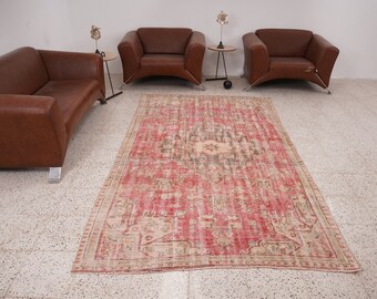 May include: A vintage rug with a faded pink and cream color scheme. The rug has a traditional Persian design with intricate details. The rug is laid out on a tiled floor in a room with brown leather furniture.