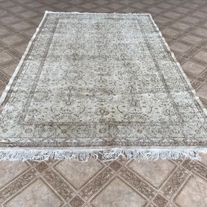 Puede incluir: Una alfombra de estilo vintage con fondo beige claro y un patrón floral marrón descolorido. La alfombra tiene un borde de flecos.