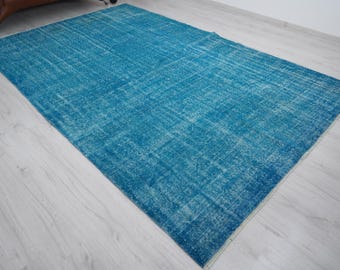 May include: A rectangular, distressed-look rug in shades of turquoise and light blue. The rug has a faded, vintage appearance with a textured surface. The rug is placed on a light-colored wooden floor.