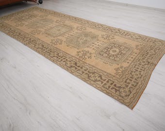 May include: A rectangular, beige-toned rug with a detailed, repeating pattern. The rug features a central design of geometric shapes and floral motifs, framed by a darker brown border. The rug is placed on a light-colored wooden floor.