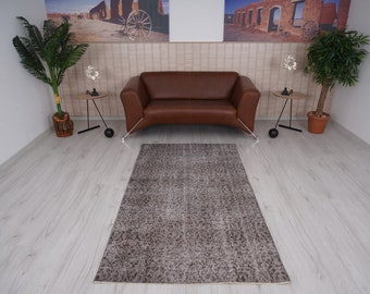 Puede incluir: Una alfombra de color marrón con un patrón de color marrón claro. La alfombra se coloca en una habitación con un sofá de cuero marrón y dos mesas auxiliares con plantas.