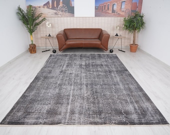 May include: A gray and white distressed area rug with a subtle pattern. The rug is laid out on a light wood floor in a room with a brown leather sofa and potted plants.