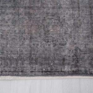 May include: A section of a gray and white patterned rug. The rug has a distressed, vintage look with a faded design. The edges are finished with a white fringe. The rug's texture is visible, with a mix of dark and light gray threads.