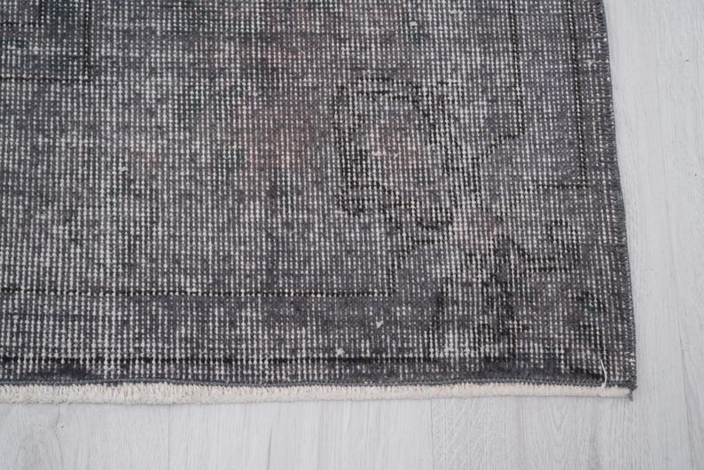 May include: A section of a gray and white patterned rug. The rug has a distressed, vintage look with a faded design. The edges are finished with a white fringe. The rug's texture is visible, with a mix of dark and light gray threads.