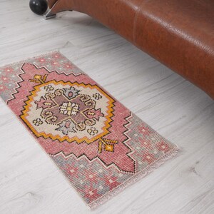 May include: A small, rectangular rug with a central medallion design. The rug features a pink and cream color palette with accents of yellow and gray. The rug is placed on a light-colored wooden floor.