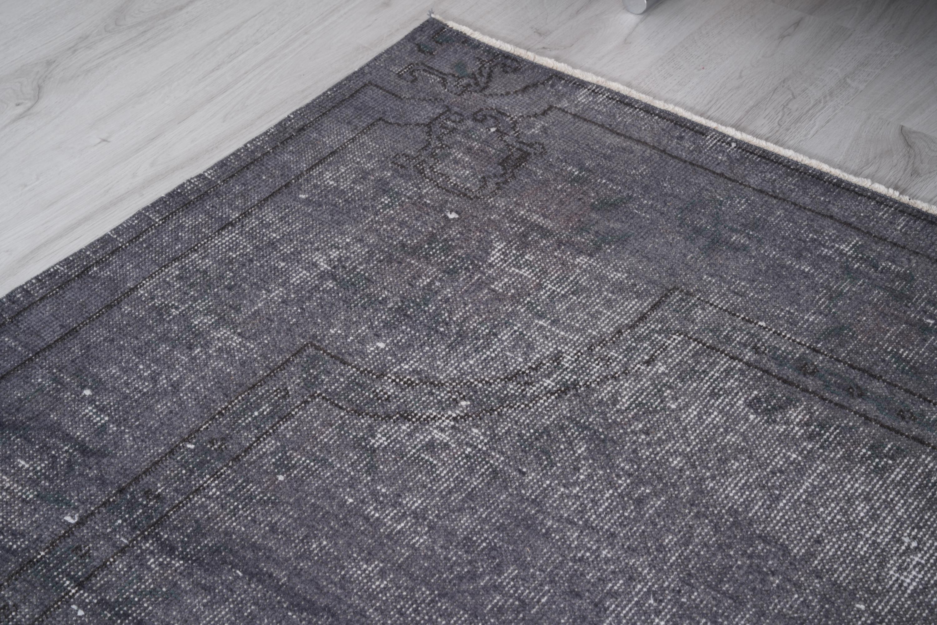 May include: A section of a dark gray vintage rug with a faded, distressed appearance. The rug has a rectangular shape with a detailed border and a subtle pattern. The rug is placed on a light gray wooden floor.