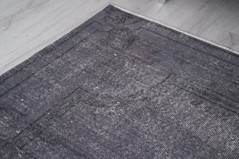 May include: A section of a dark gray vintage rug with a faded, distressed appearance. The rug has a rectangular shape with a detailed border and a subtle pattern. The rug is placed on a light gray wooden floor.