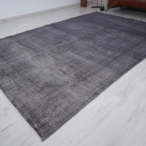 May include: A large, rectangular area rug in shades of gray and charcoal. The rug has a distressed, vintage appearance with a faded pattern and a white border. The rug is placed on a light-colored wooden floor.