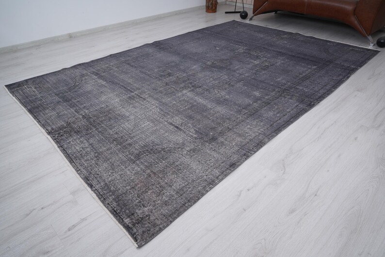 May include: A large, rectangular area rug in shades of gray and charcoal. The rug has a distressed, vintage appearance with a faded pattern and a white border. The rug is placed on a light-colored wooden floor.