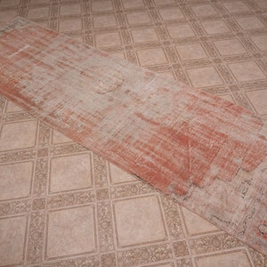 May include: A long, rectangular rug with a faded, distressed look. The rug has a light beige background with a subtle, faded red pattern. The rug is lying on a tiled floor.