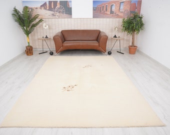 Può includere: Un grande tappeto rettangolare color crema con un sottile motivo floreale nei toni del marrone e del beige. Il tappeto è posato su un pavimento in legno chiaro, con un divano in pelle marrone e tavolini sullo sfondo. La stanza è ben illuminata.