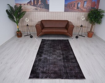 Puede incluir: Una alfombra vintage gris oscuro con un aspecto desgastado, colocada en un salón con un sofá de cuero marrón y dos mesas auxiliares con detalles decorativos.