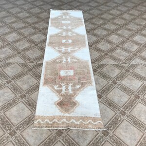 May include: A white and brown runner rug with a geometric pattern. The rug has a faded look and is made of wool.