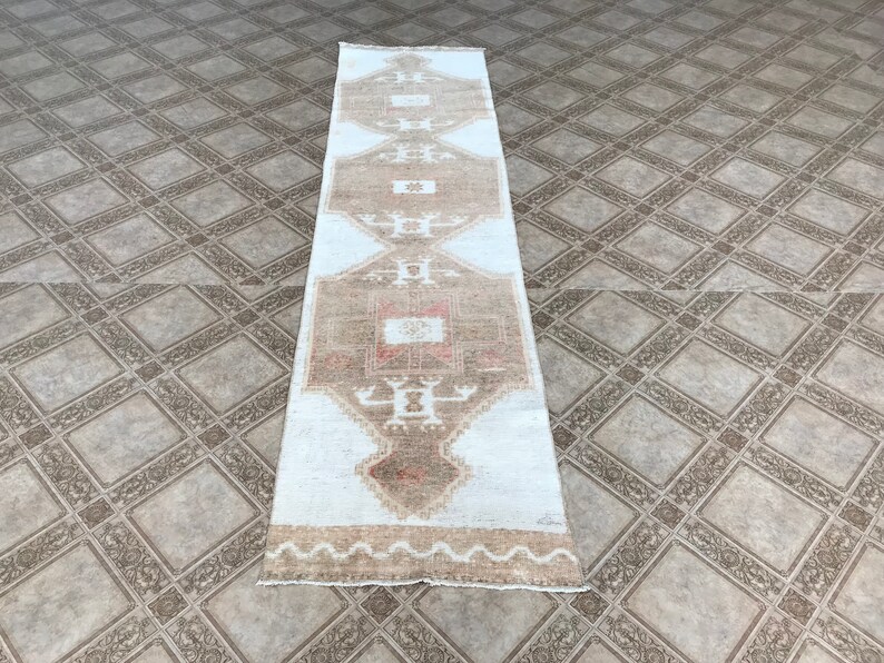 May include: A white and brown runner rug with a geometric pattern. The rug has a faded look and is made of wool.