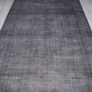May include: A rectangular, distressed gray rug with a faded appearance. The rug has a border and a textured surface, with variations in the gray tones. The rug is placed on a light-colored wooden floor.