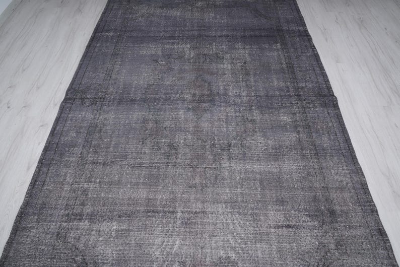May include: A rectangular, distressed gray rug with a faded appearance. The rug has a border and a textured surface, with variations in the gray tones. The rug is placed on a light-colored wooden floor.