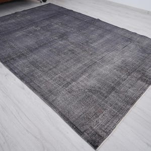 May include: A rectangular, distressed-look rug in shades of gray and black. The rug has a faded appearance with visible texture and a subtle pattern. The rug is placed on a light-colored wooden floor.