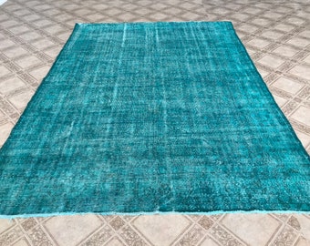 May include: A large, rectangular rug in shades of teal and turquoise. The rug has a distressed, vintage appearance with a faded, textured pattern. The edges are slightly frayed, adding to the rug's aged aesthetic. The rug is placed on a patterned tile floor.