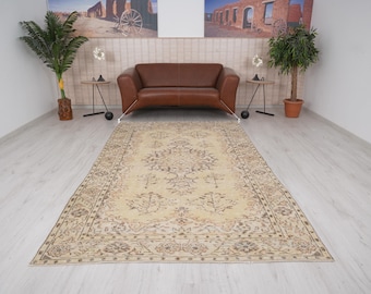 Op de afbeelding: Een vintage crèmekleurig tapijt met een bloemenpatroon en een bruine rand. Het tapijt ligt op een lichtgrijze houten vloer in een kamer met een bruine leren bank en twee planten in potten.