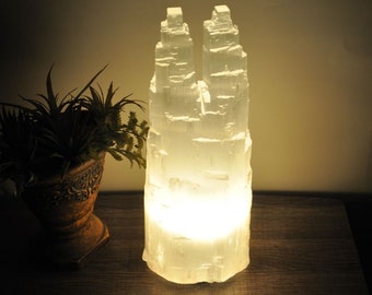 May include: A white selenite crystal lamp with a warm glow. The lamp is shaped like a tower and has a natural, rough texture. The lamp is sitting on a wooden surface.