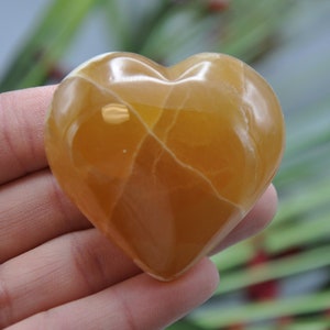 May include: A polished, heart-shaped stone with a warm, amber hue. The stone has natural white veining throughout, adding to its unique character. The heart is held in a hand, with a blurred background of greenery.
