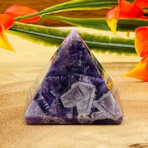 May include: A purple amethyst pyramid crystal with a smooth, polished surface. The pyramid is sitting on a wooden surface with orange flowers in the background.