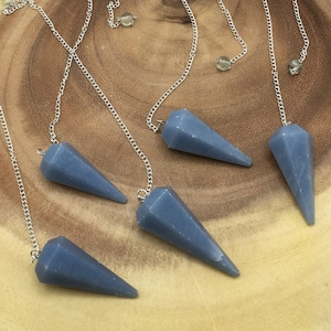 May include: Five blue crystal pendulums with silver chains and small crystal beads. The pendulums are cone-shaped and lie on a light brown wooden surface. The chains are attached to the top of each pendulum.
